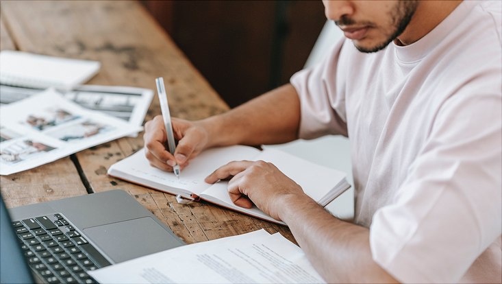 Ein Mann sitzt an seinem Laptop und schreibt auf einem Block Papier.