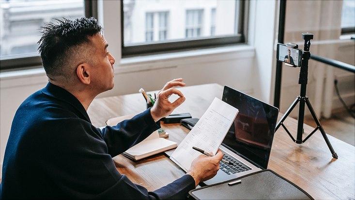 Eine Mann sitzt an seinem Laptop und nimmt sich selbst mit dem Handy auf.