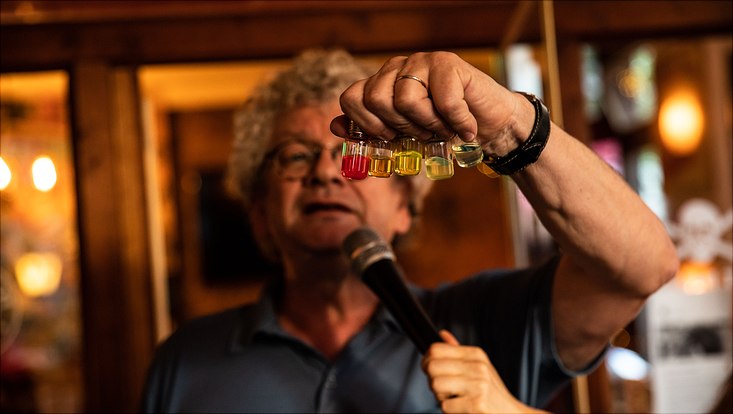 Ein Mann hält kleine Gefäße mit unterschiedlichen Flüssigkeiten hoch. Gleichzeitig hält ihm jemand ein Mikrofon vor.