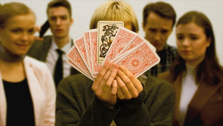 Im Hintergrund sind unscharf fünf Personen zu sehen, eine der Personen in der Mitte hält neun Karten in den Vordergrund. Die Karten sind scharf zu sehen, eine davon zeigt ein Skelett, die restlichen sind umgedreht.