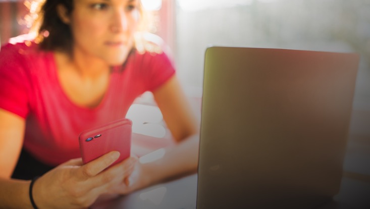 Eine Frau sitzt vor einem Laptop und hält ein Handy in der Hand