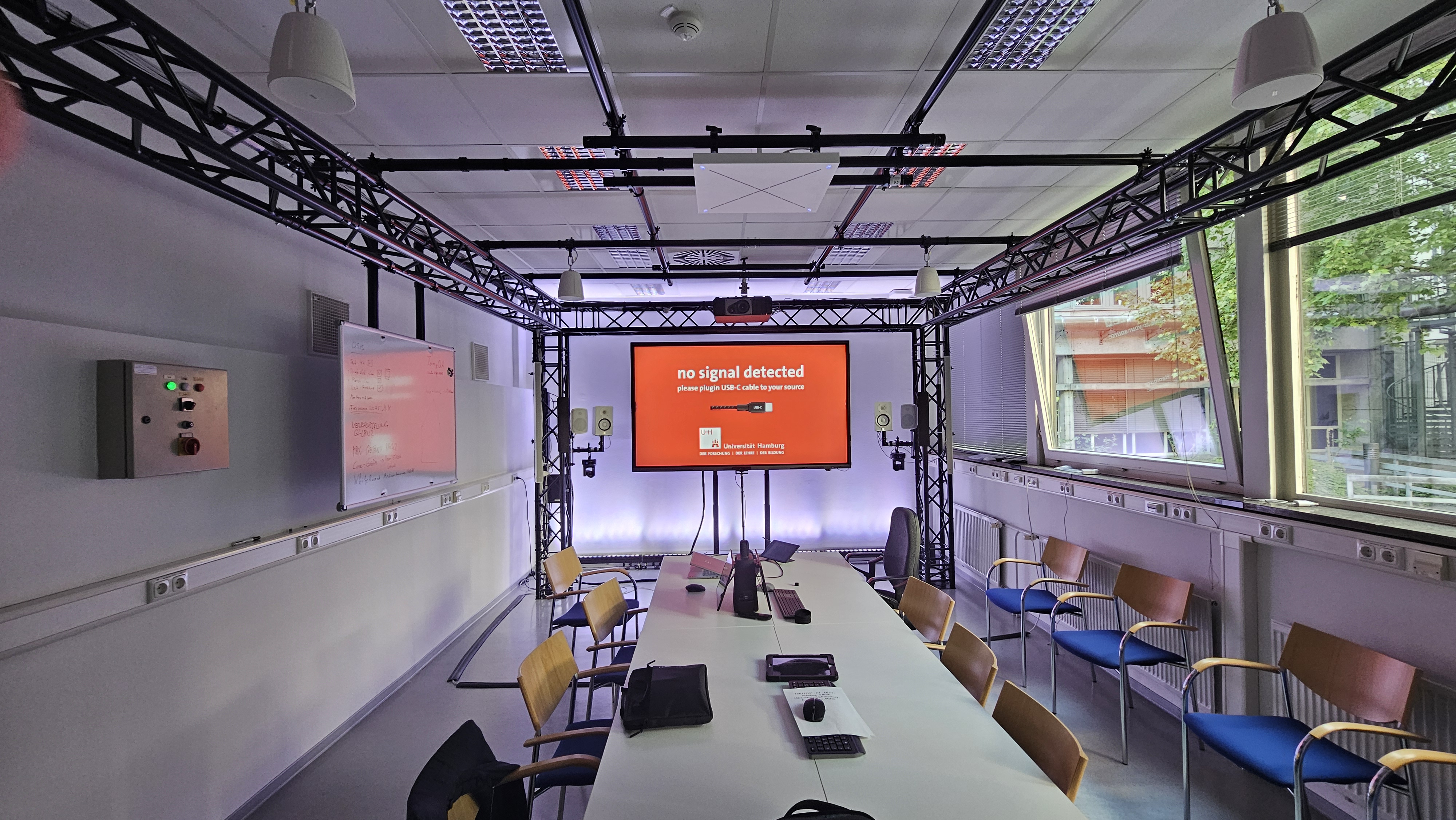 Ein modern ausgestatteter Seminarraum mit einem großen Konferenztisch, Stühlen und einem großen Bildschirm, auf dem „No signal detected“ steht. Um den Raum verläuft eine Metallkonstruktion mit montierten Lautsprechern, Kameras und Lichttechnik.