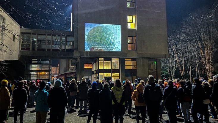 Es ist dunkel, mehrere Menschen stehen vor einem Gebäude und schauen einen Film, der an die Hauswand projiziert wird.